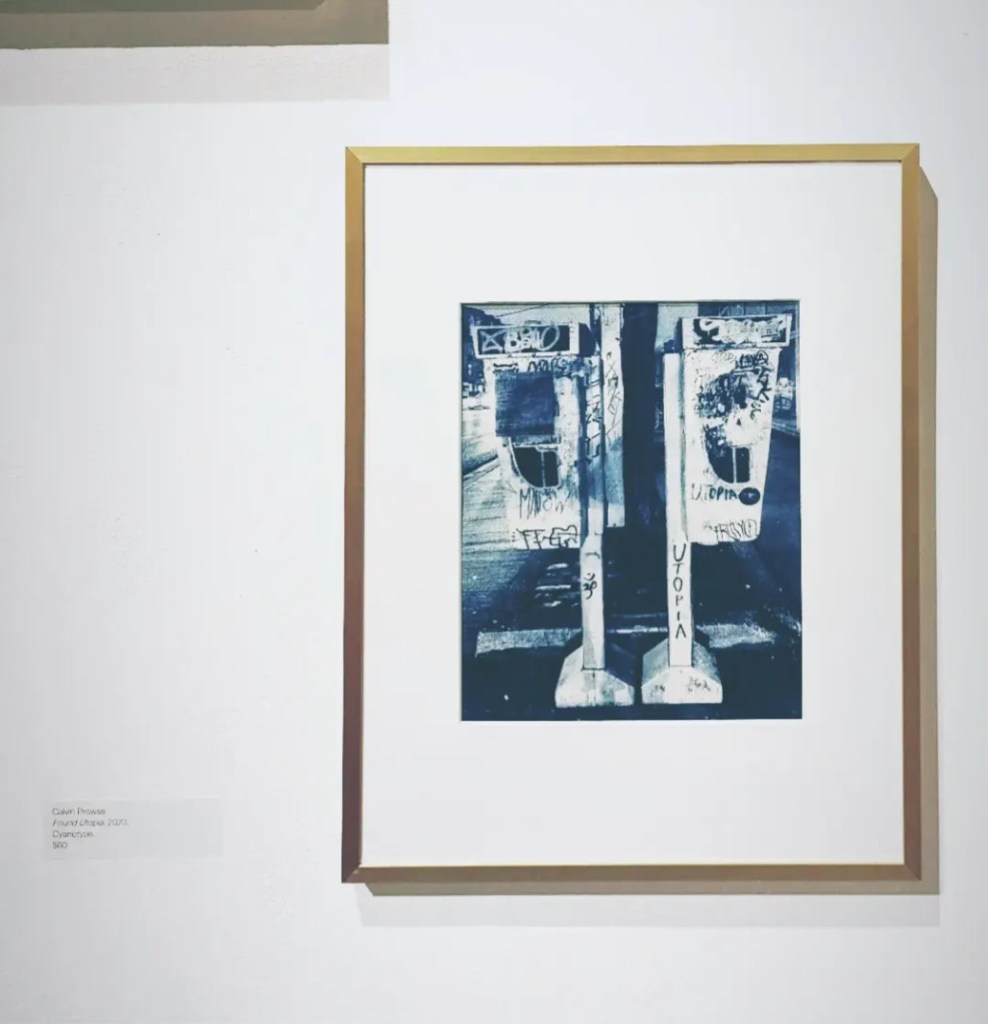 A blue photograph of two payphones side by side, with the word utopia written on them in sharpie, in a gold frame. 